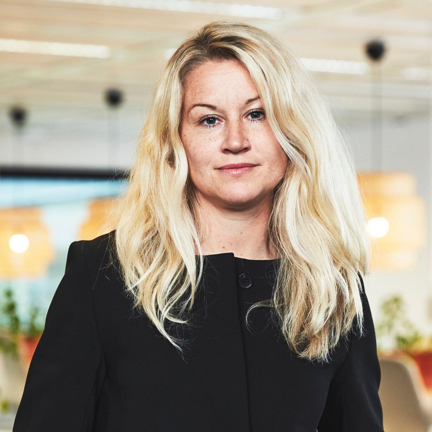 A portrait of a blond woman in a black short, standing in an office environment.