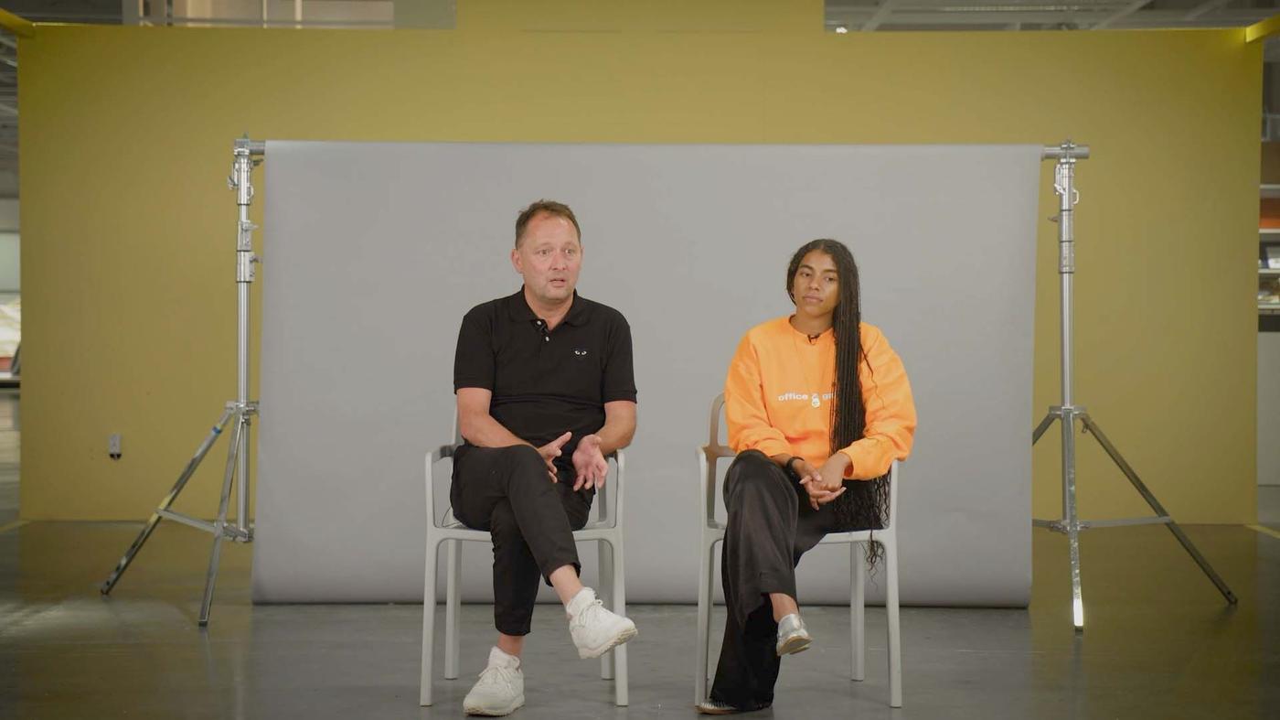 A video of a man and a woman talking they are sitting in front of a grey backdrop and a yellow wall.