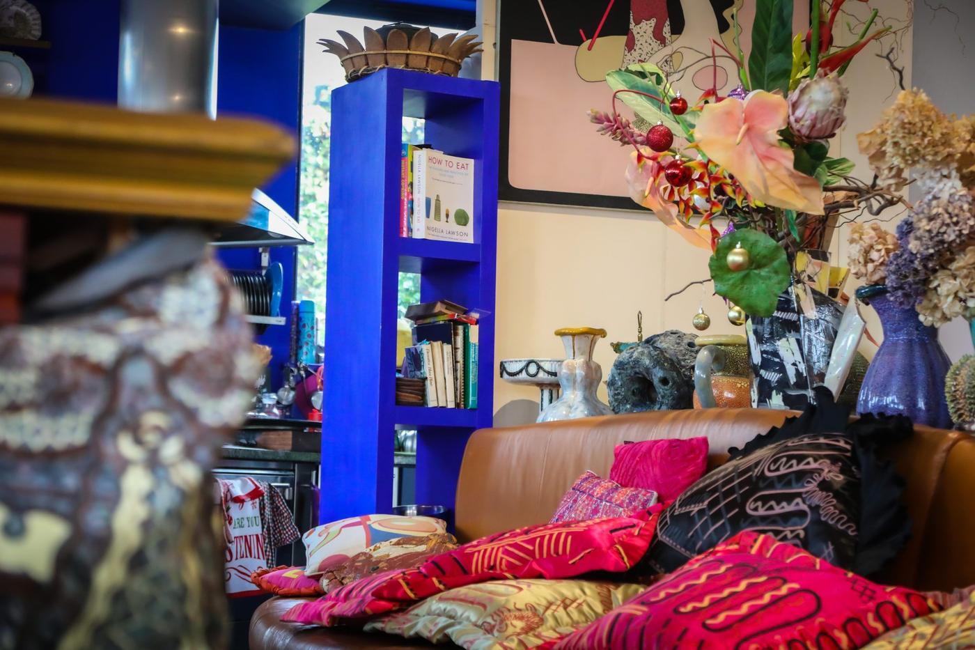An eclectic composition of flowers, vases, cushions and books, a bright blue bookshelf and a brown couch.