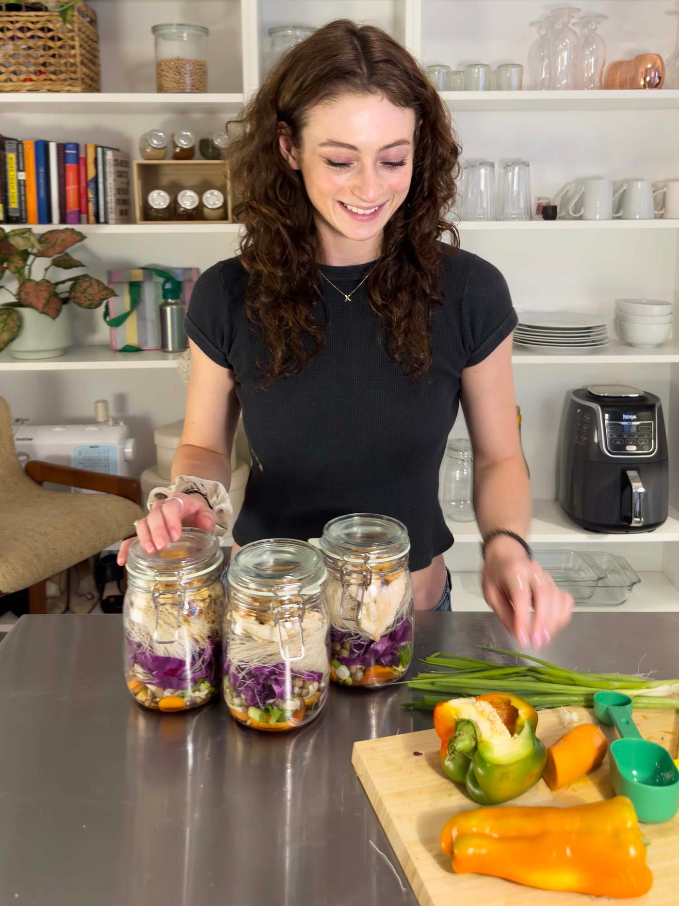 Leah stands at the worktop. She looks down at her ingredients and ramen soup jars.