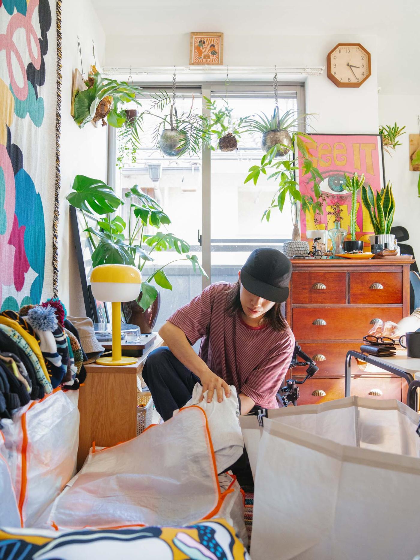 Daisuke packs clothes into a transparent storage bag. His room is full of plants and personal belongings.