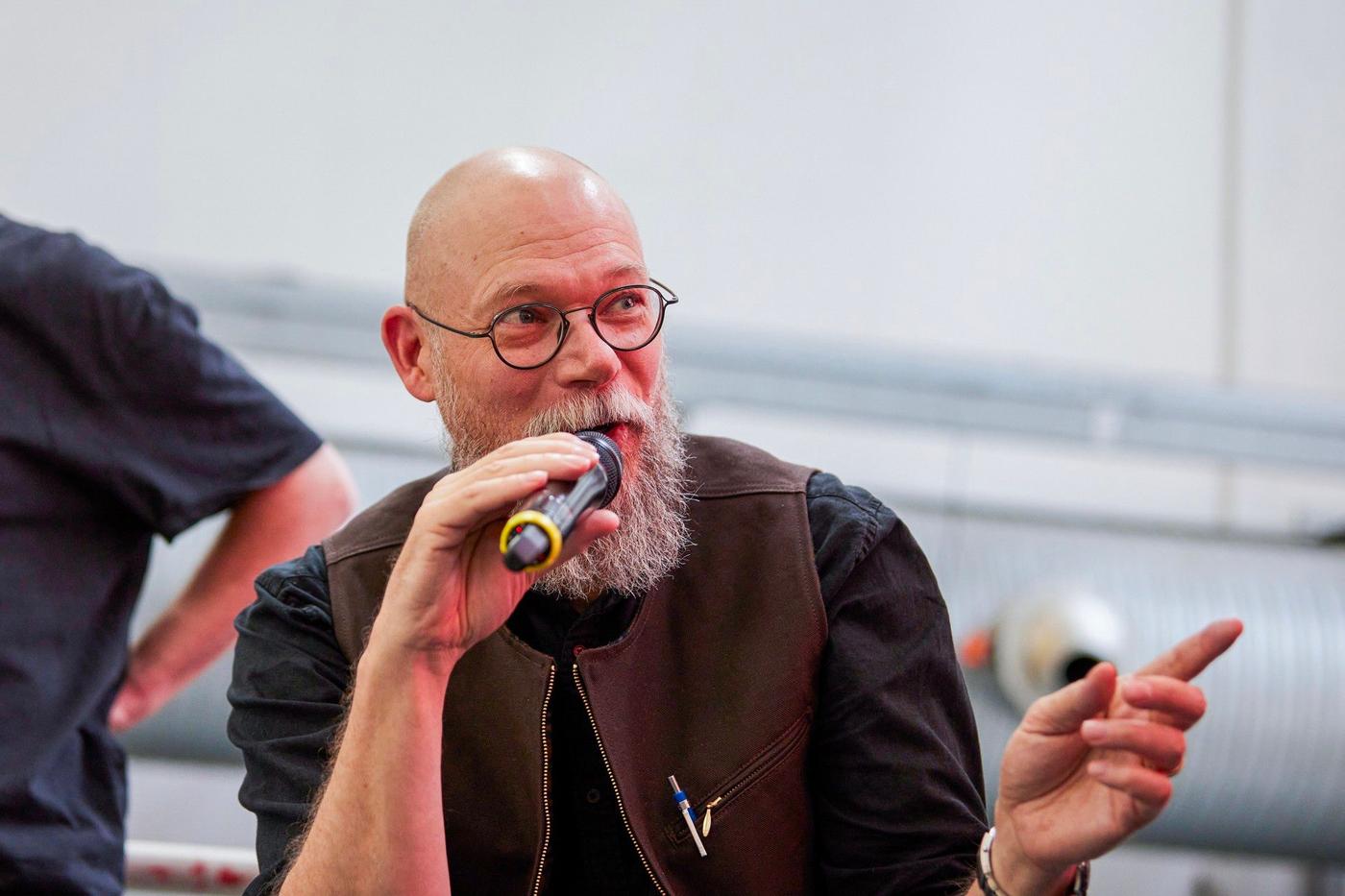 A bald man with a beard in a brown vest sitting on a stage with a microphone in his hand.