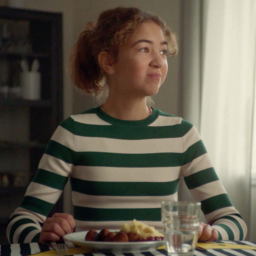 A girl in a striped top sits at the table. She is looking out of the window, while eating a plate of meatballs and mashed potato.
