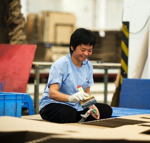 In a factory environment, a woman is sitting with a brush in her right hand and a glove in the left hand.