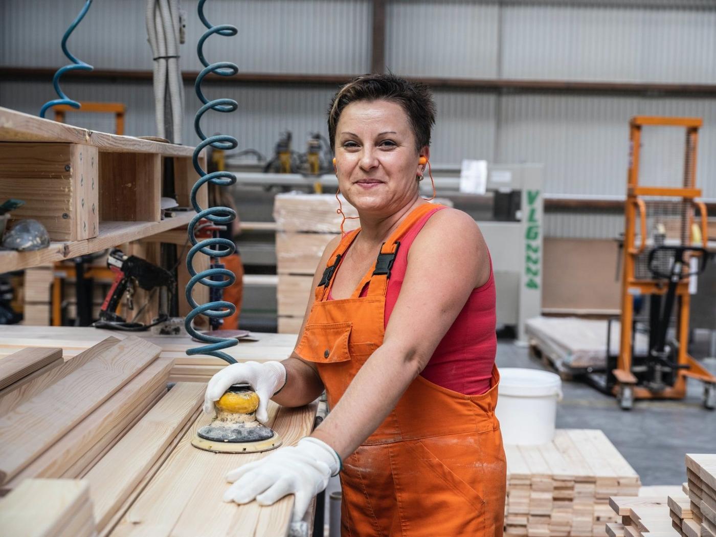 A woman in orange work ware standing in an industry environment sanding a plank.