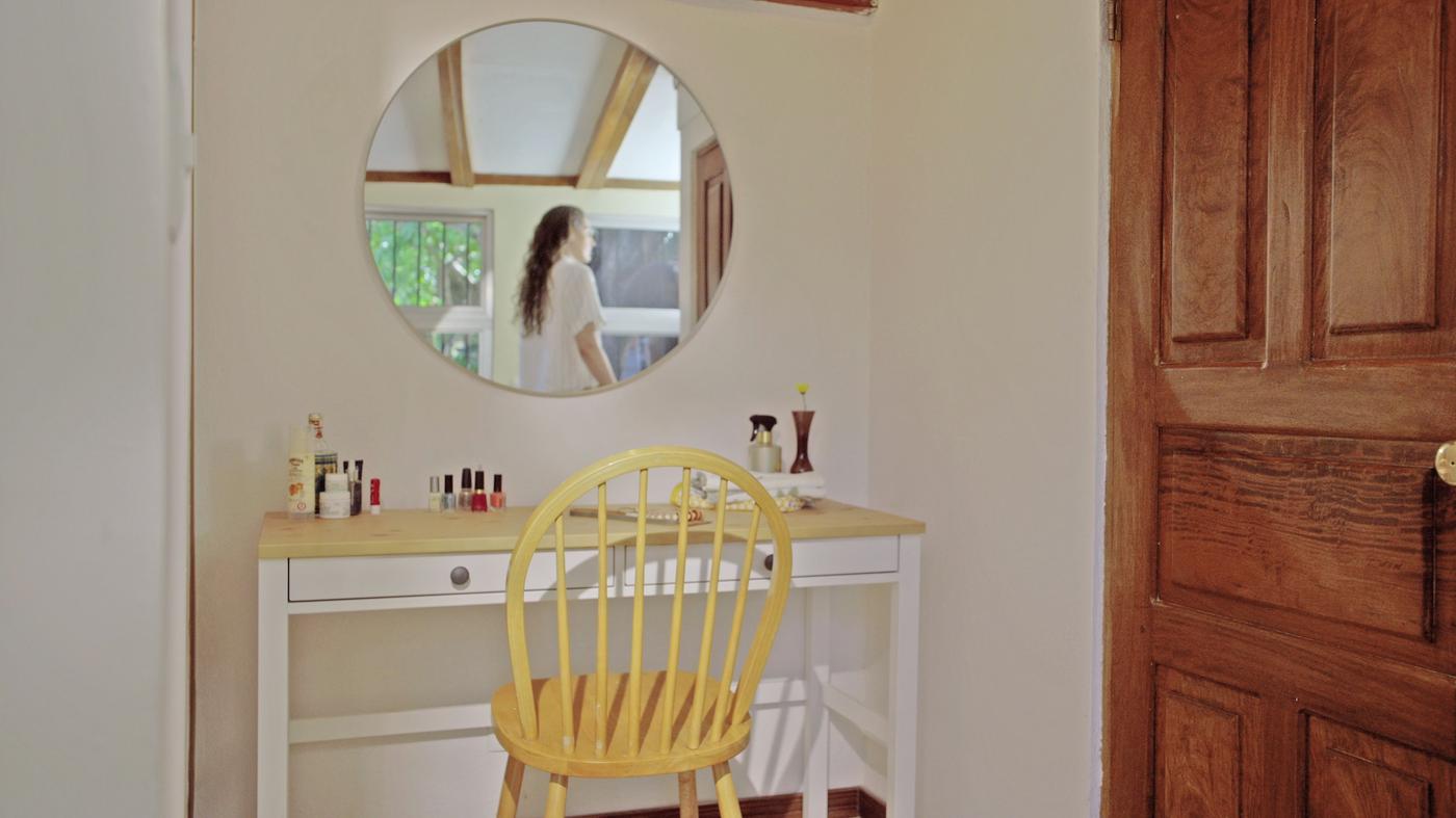 In the bedroom, make up and brushes are arranged on a dressing table, with a yellow chair in front and a round mirror above.