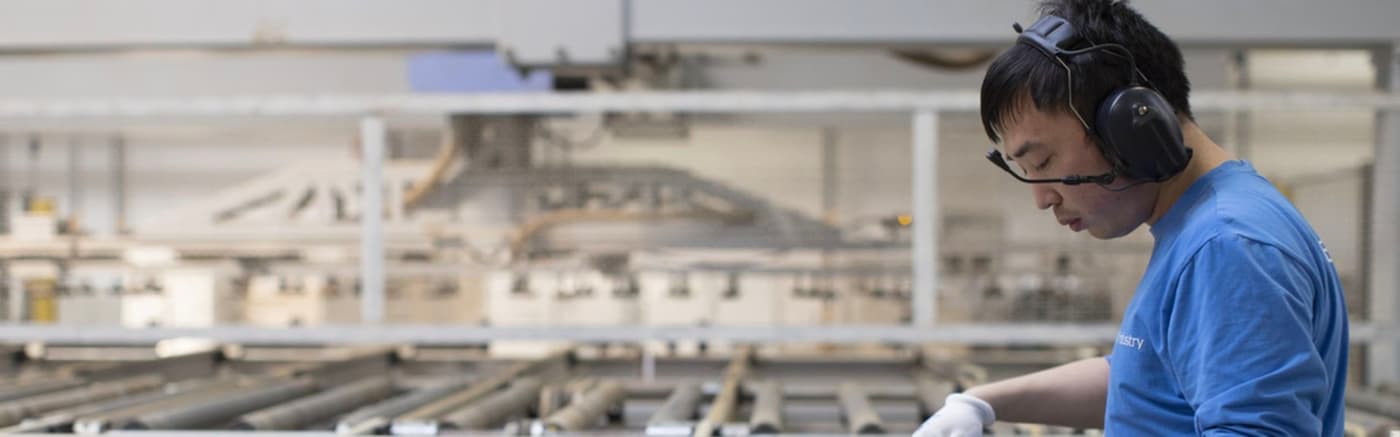 A man in a blue shirt standing in an industrial environment measuring a board.
