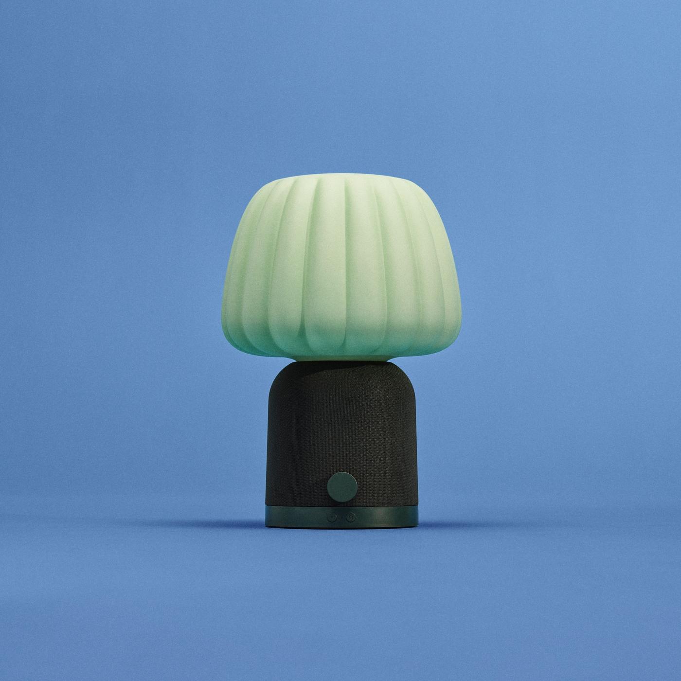 Dark green KULGLASS speaker with mesh-textured base and lighter pistachio-toned patterned shade, resembling a table lamp, set against a blue background.