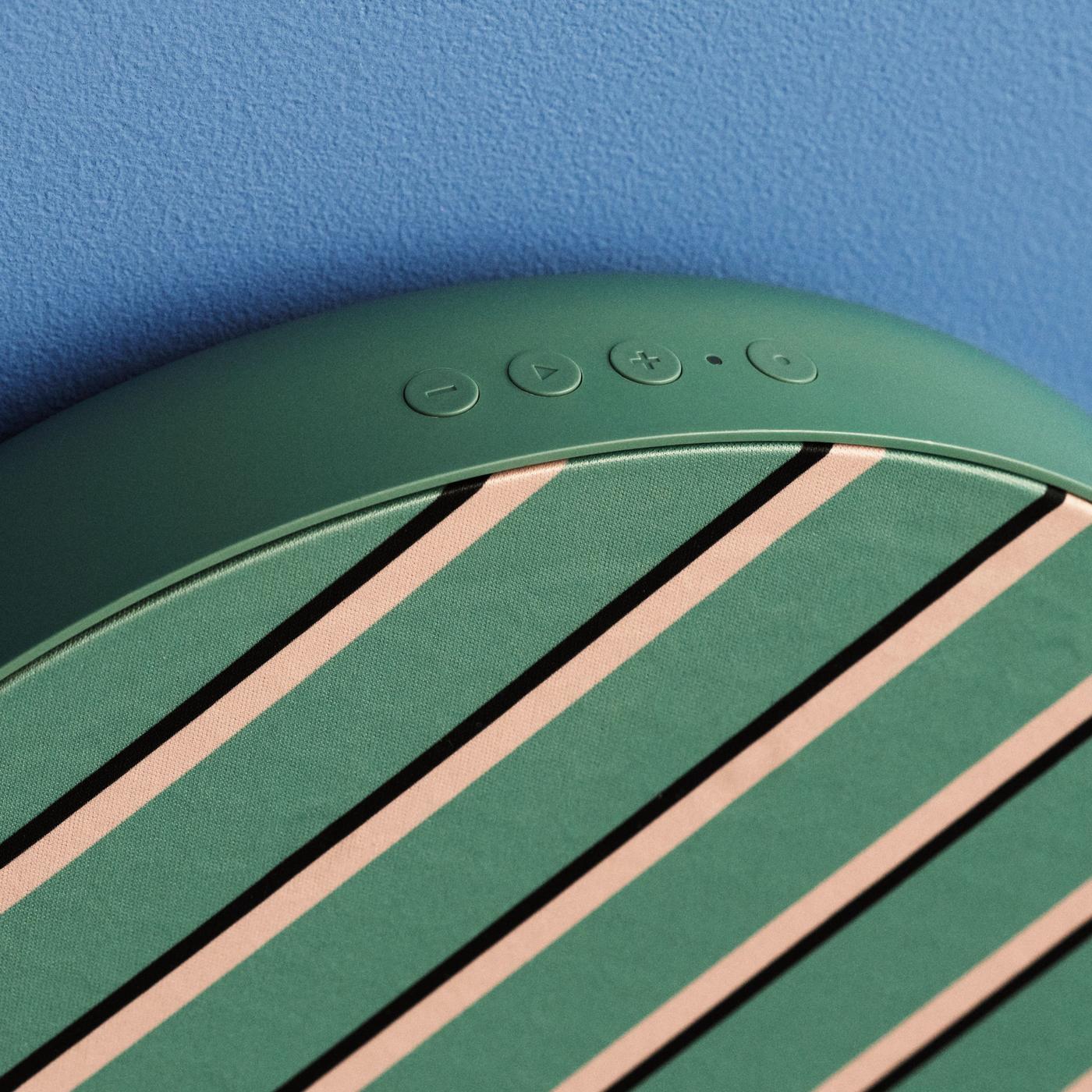 Close-up of the top of green patterned surface of a SOLSKYDD speaker, showing control buttons for play and volume, with detailed view of the patterned fabric and blue background