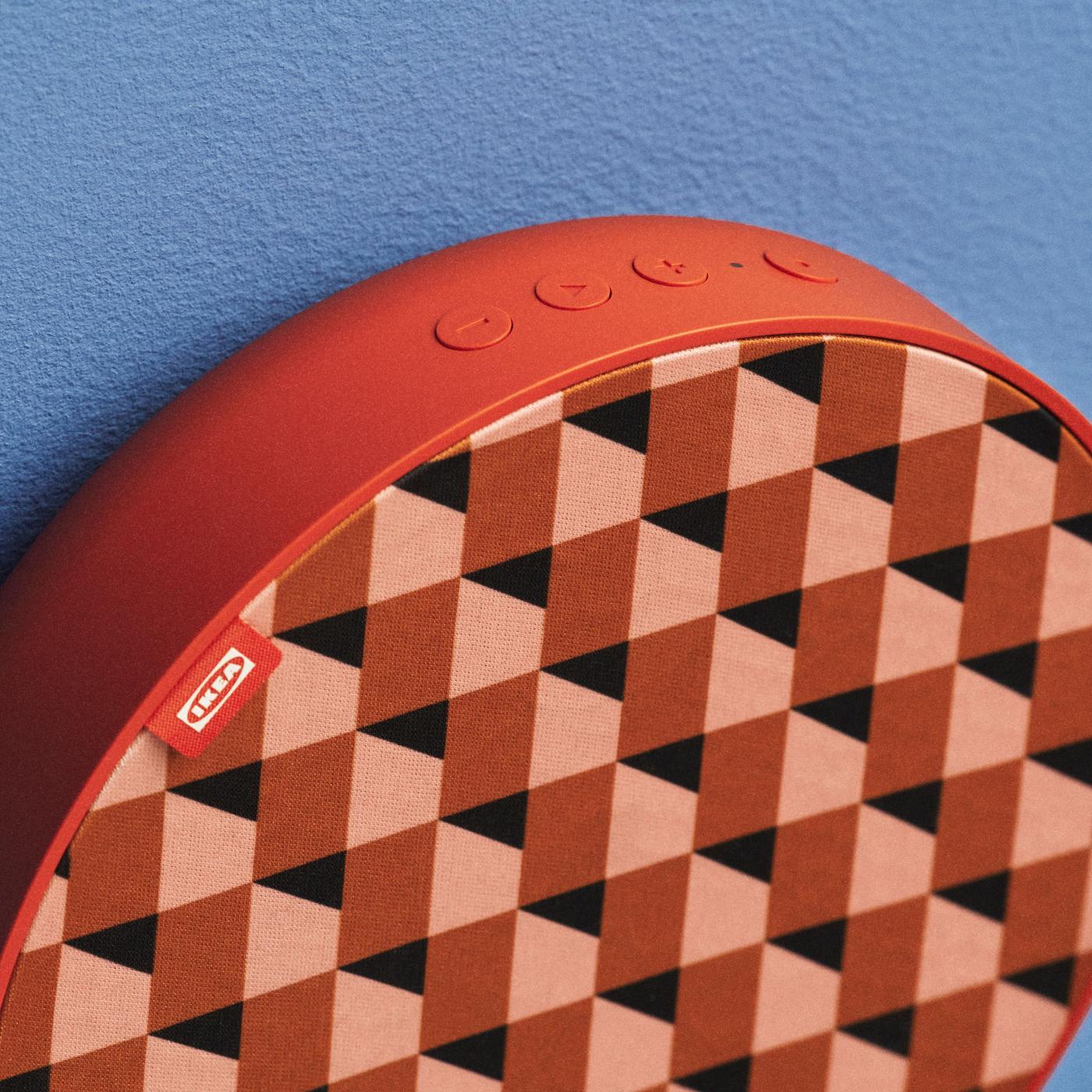 Close-up of a red round SOLKSYDD speaker with red-pink zigzag pattern, showing top-side control buttons, set against a blue background.