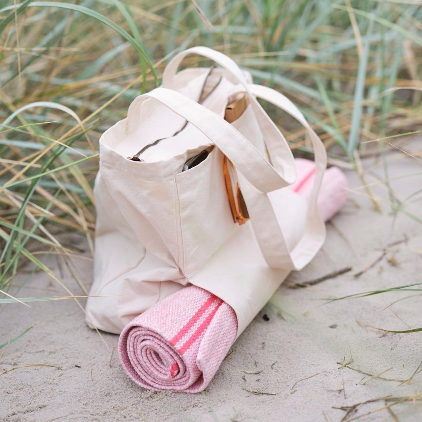 A strand bag packed with a pink yoga rug standing at the beach.