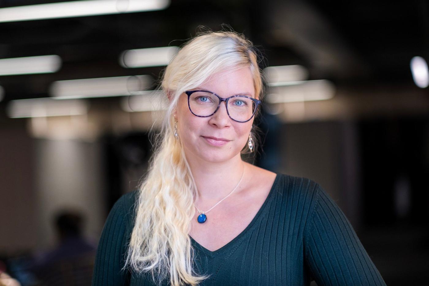 A blonde woman with glasses standing in a office environment.