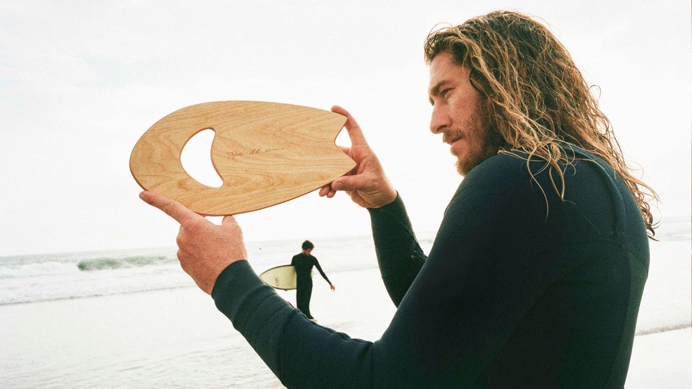 A surfer in a wet suite standing at the beach holding a hand pane