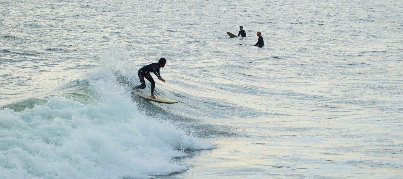 A single surfer catches an evening wave. Nearby, two other surfers are sitting on their boards waiting for a wave.