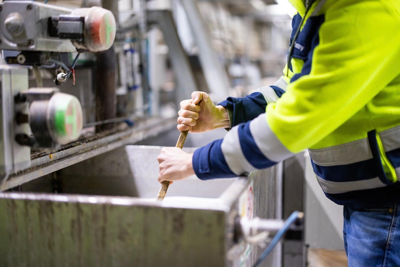 An industrial worker in a yellow jacket stirring a metal container with glue.