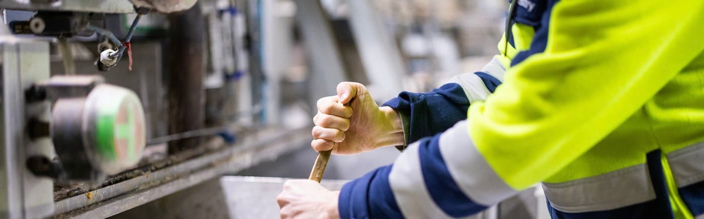 A factory worker in a yellow jacket stirring with a wooden stick in a metal container.