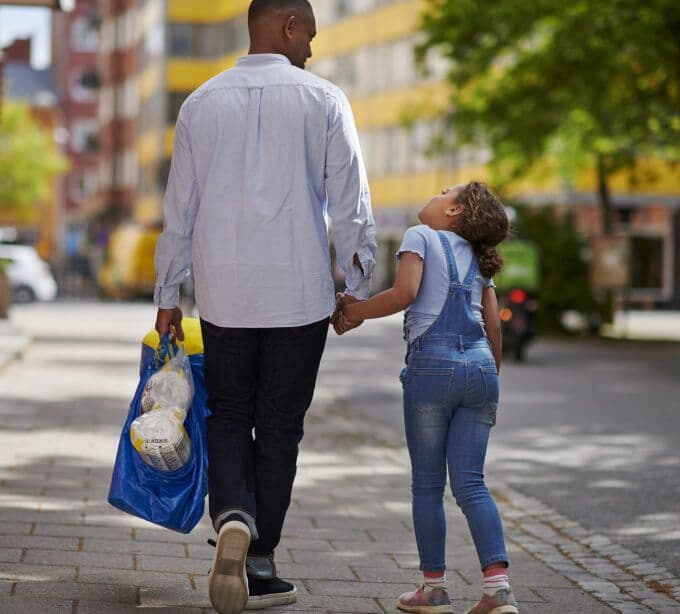 A parent and child walking hand in hand down a street; the parent holding a blue FRAKTA bag filled with IKEA products.