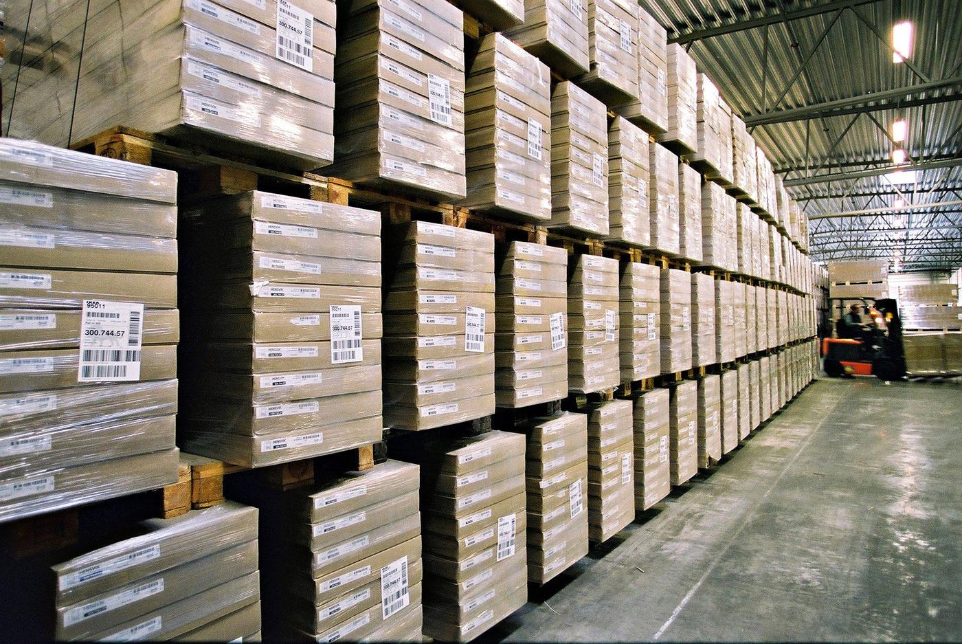 Identically packaged and stacked pallets form a high wall in a warehouse. A forklift operates nearby.