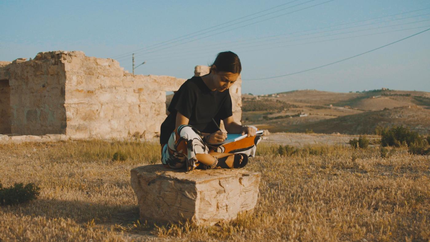 A woman sitting cross-legged on a stone under a blue sky in a dry landscape.