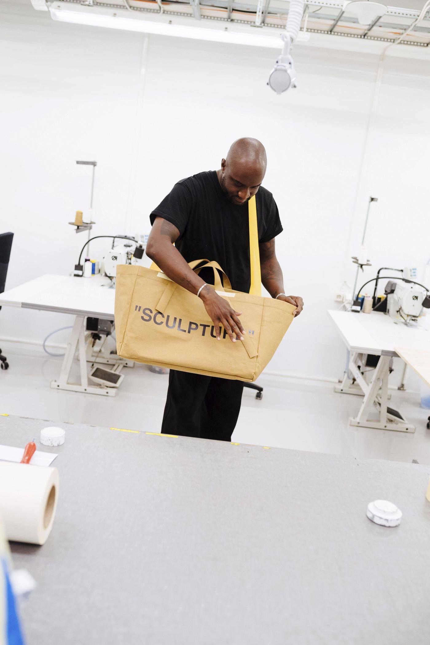 A man standing in a studio with a yellow carrier bag slung over his shoulder.