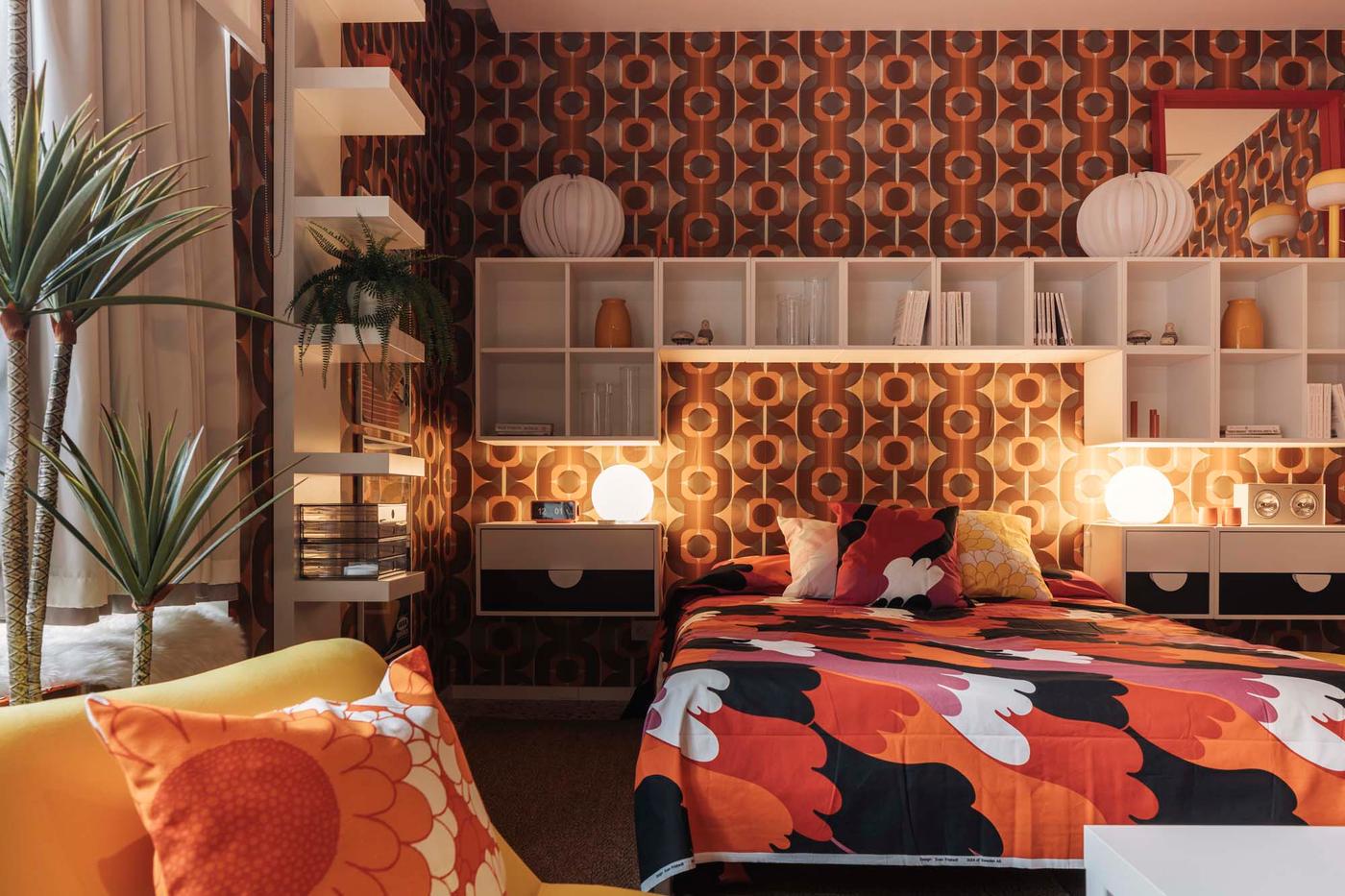 A bedroom with retro wallpaper and a white shelving unit above a bed dressed in bedding made from SVEDJENÄVA pre-cut fabric.