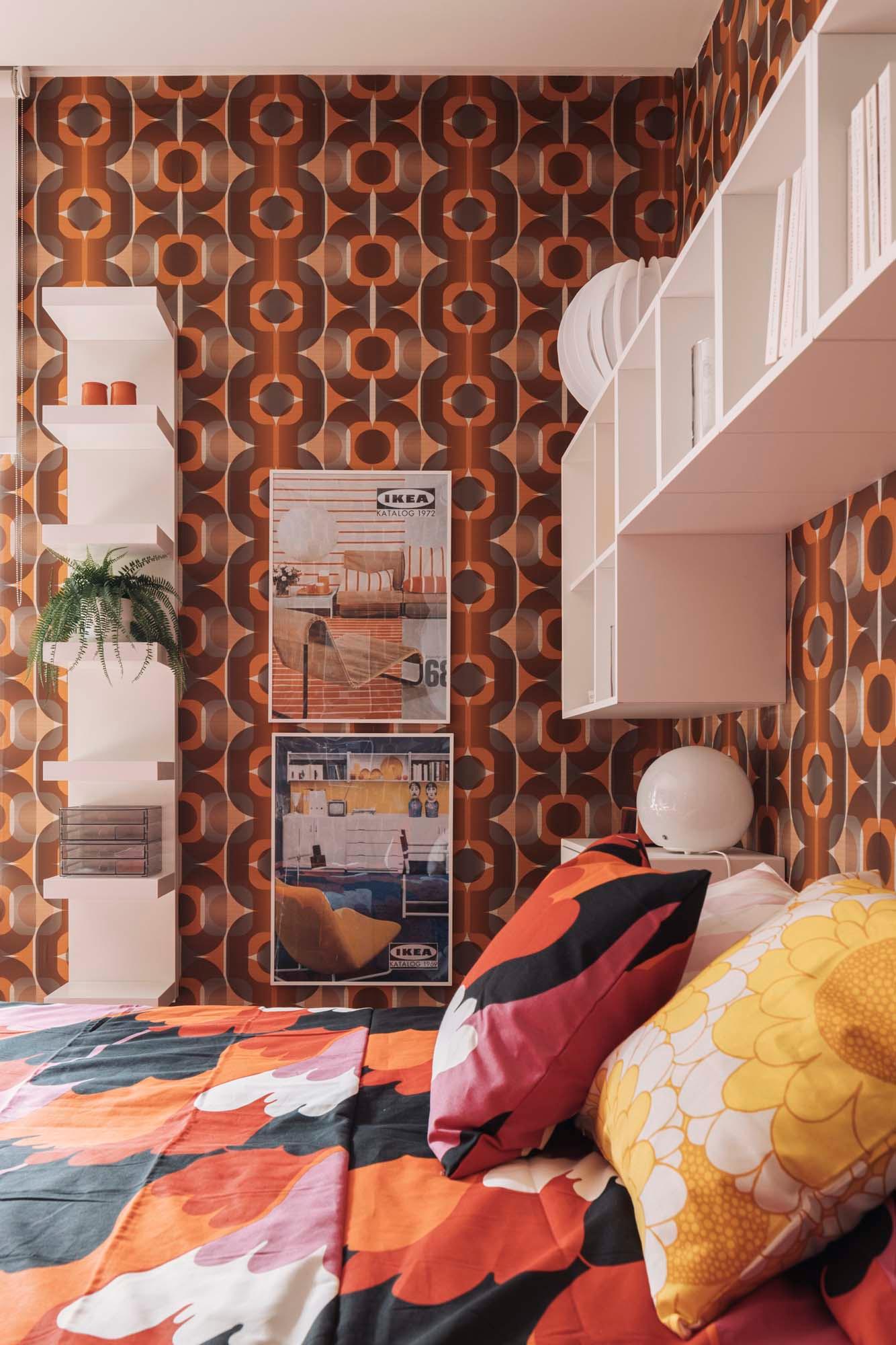 A bedroom with retro wallpaper and a white shelving unit above a bed dressed in bedding made from SVEDJENÄVA pre-cut fabric.