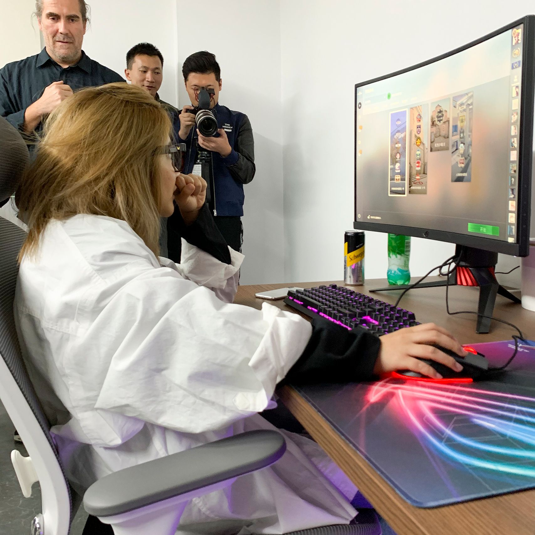 A woman in a white shirt sitting on a grey/white office chair at a desk with a computer; three men are in the background.