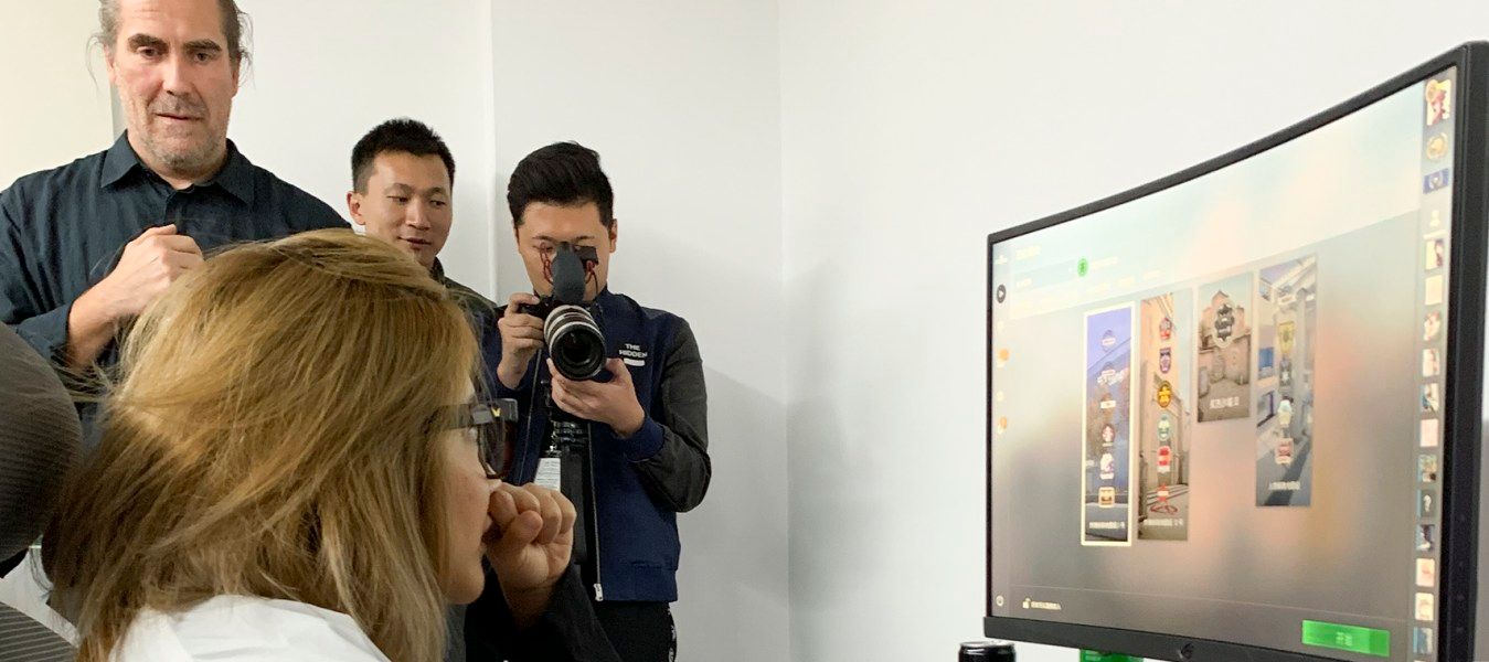 A woman in a white shirt sitting on a grey/white office chair at a desk with a computer; three men are in the background.