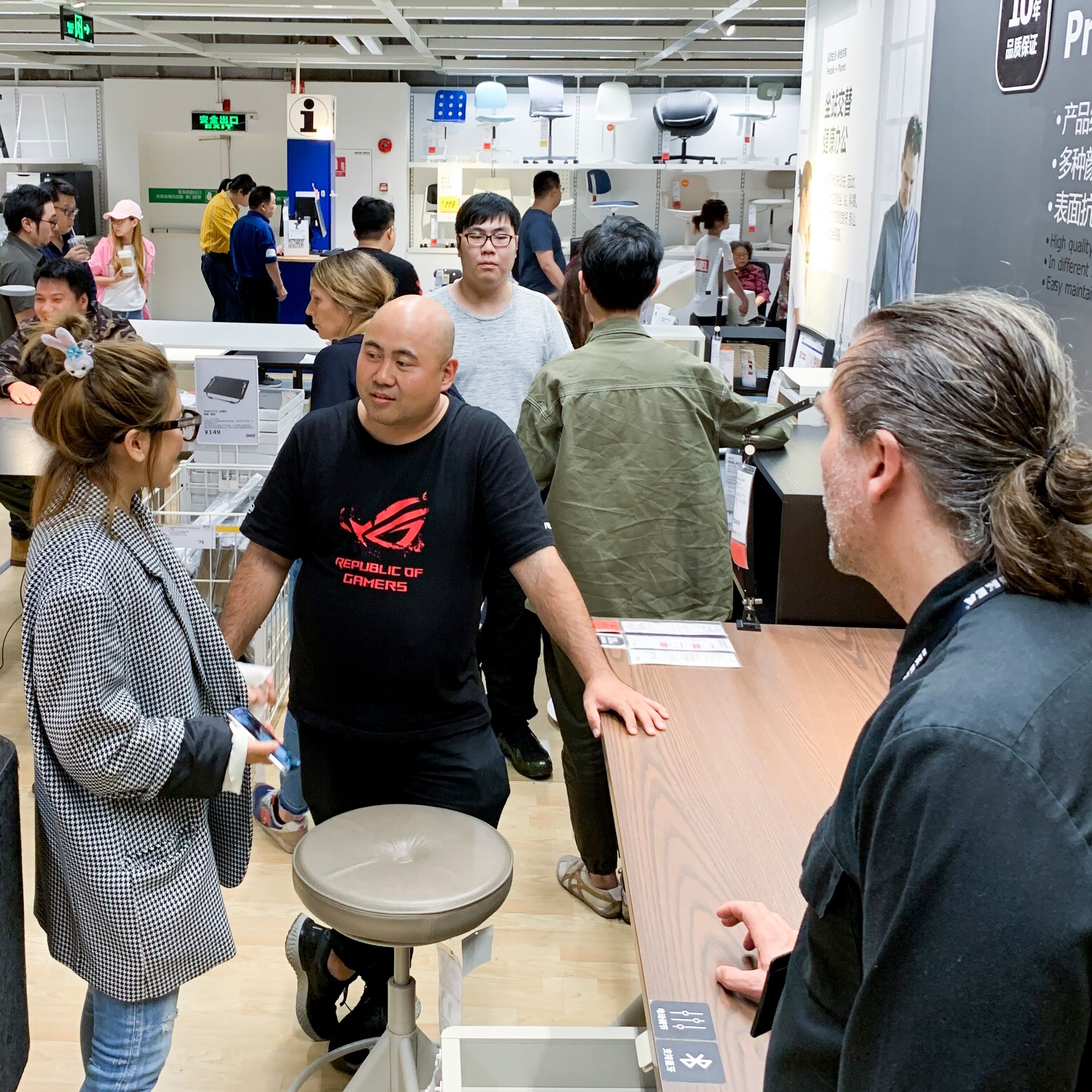 People engaged in conversation among desks inside an IKEA store, with swivel chairs on white shelving in the background.