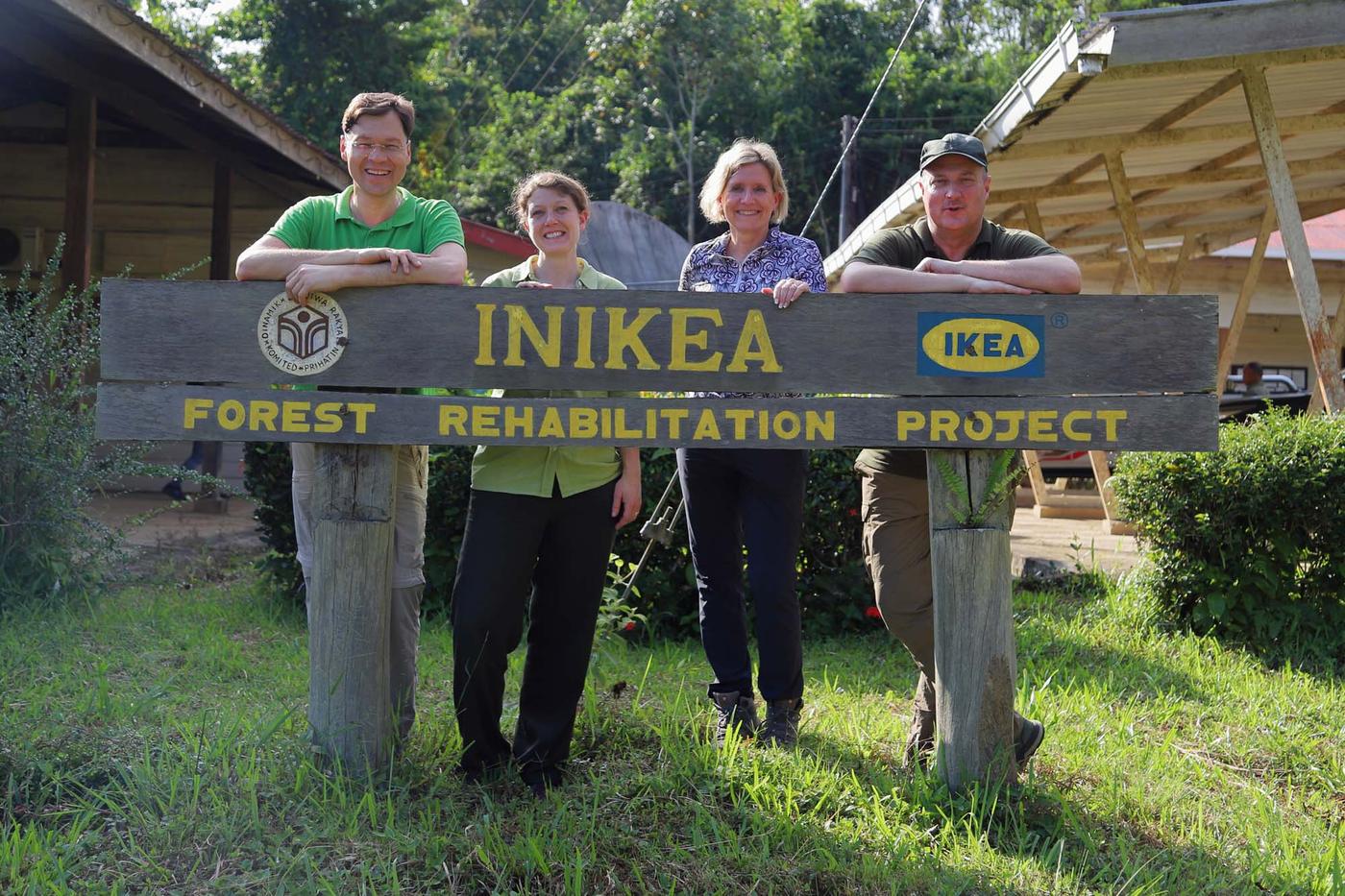 Four people stand behind a wooden sign for "INIKEA forest rehabilitation project". They stand outside, on the grass, in front of trees.