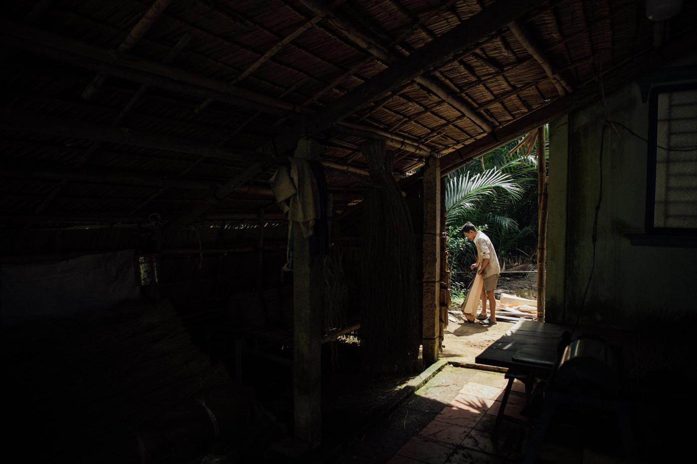 A man working a tree trunk with a machete in front of the sunny entrance of a house.