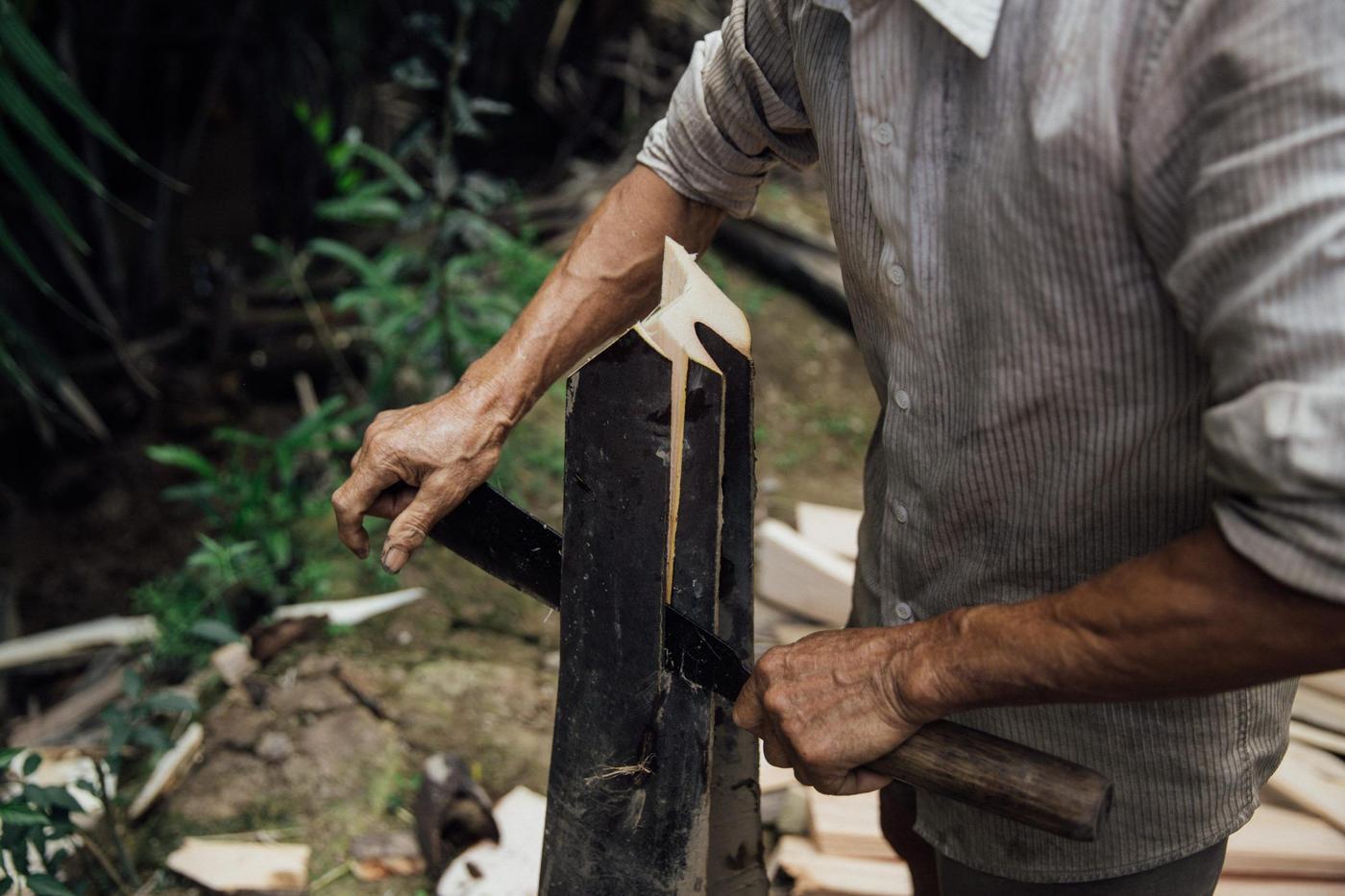 A man with a dirty shirt and sunburnt arms cutting a tree trunk vertically with a machete.