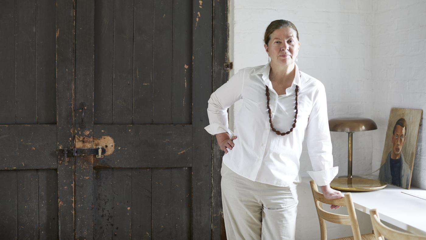 Ilse Crawford stands leaning with one hand on the back of a dining chair and the other on her hip and she looks at the camera.