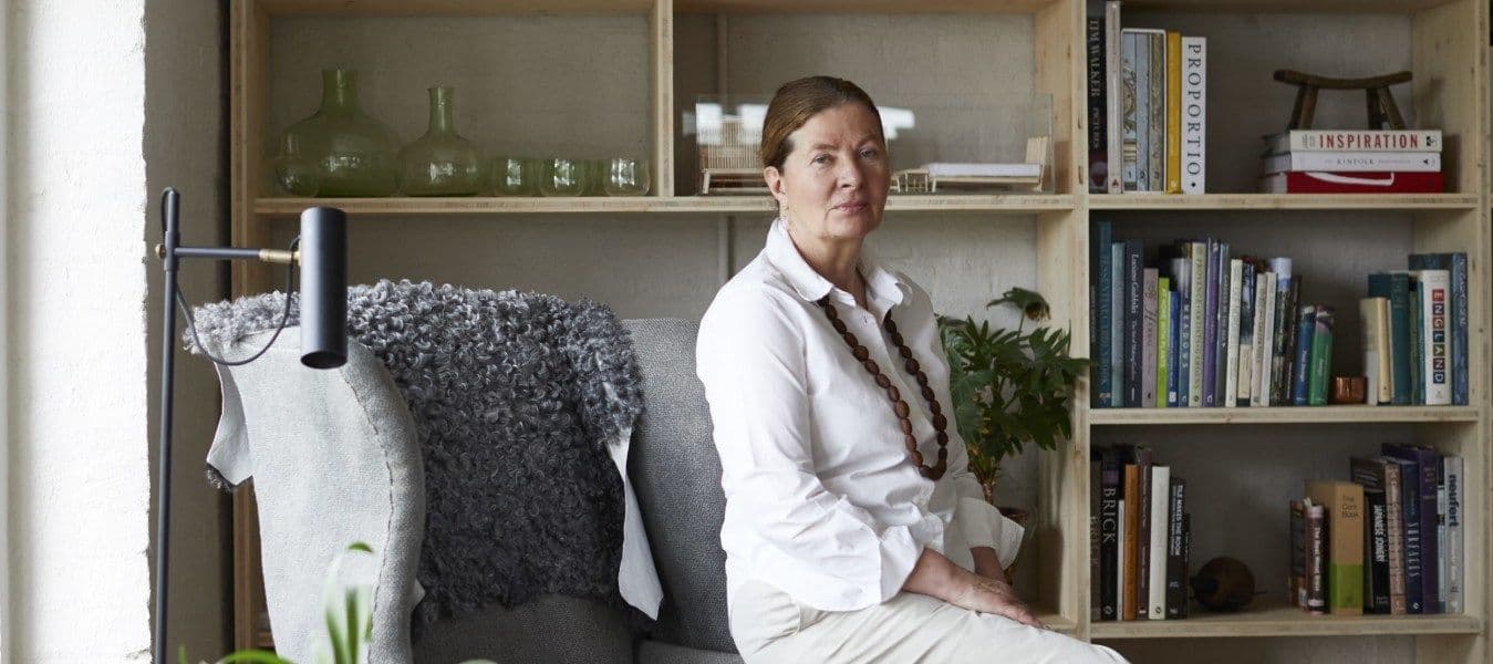 A portrait of designer Ilse Crawford, sitting on the armrest of a grey armchair, surrounded by books, decorations and plants.