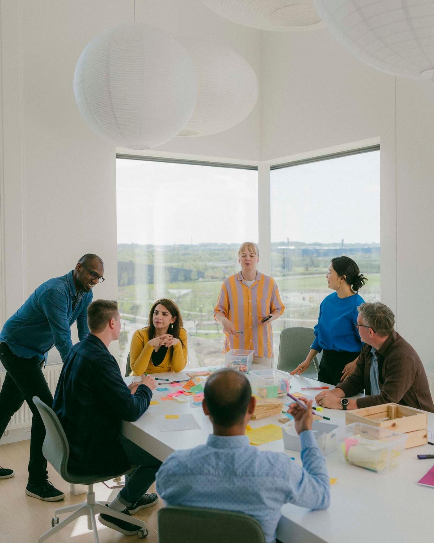 A group of seven people stand and sit in a meeting room. They are gathered around a long table. There is a corner window in the background behind them.