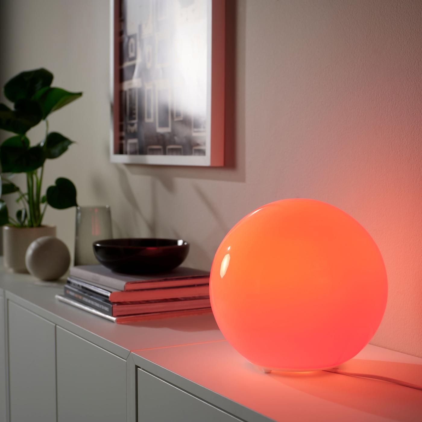 A glowing spherical table lamp lit in deep red, placed on a white shelf with artwork, books, and plants in the background.