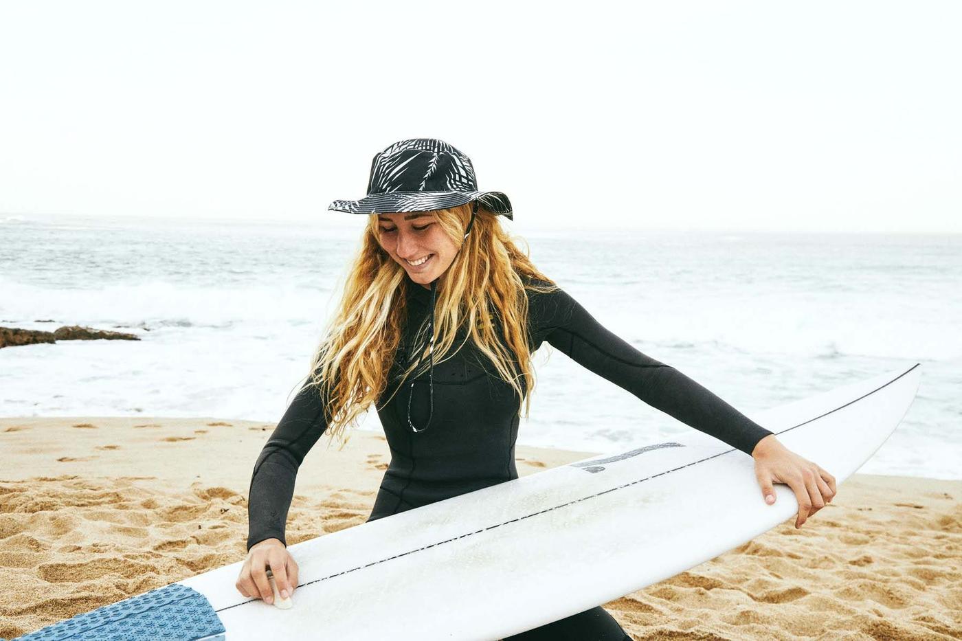 Surfer wearing a hat and wetsuit holds a surfboard on a beach.