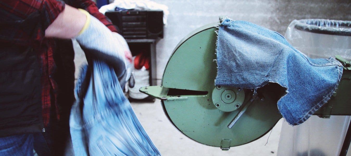 A person wearing grey gloves putting jeans into a green machine with a piece of denim fabric on top.