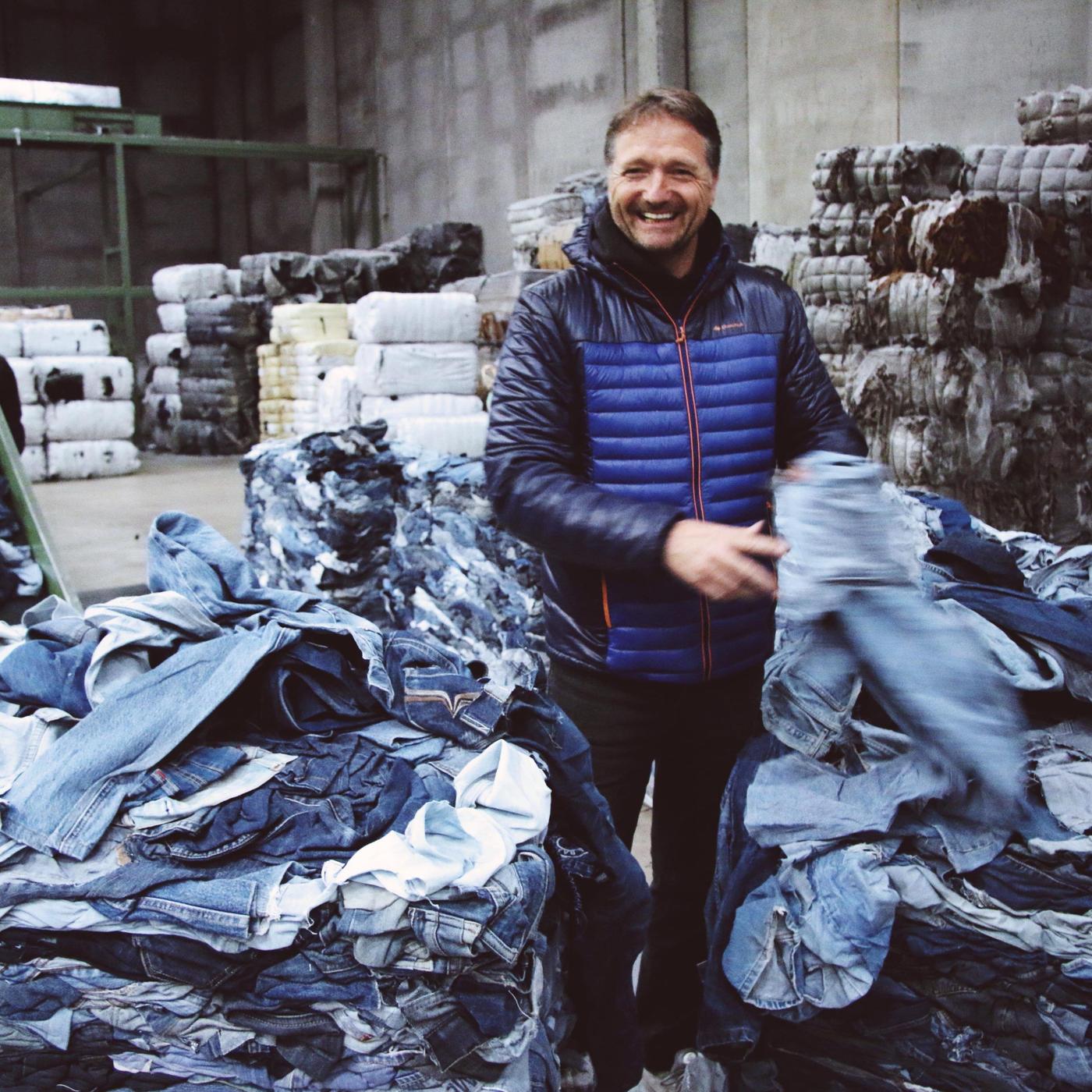 A man in a blue jacket surrounded by piles of worn-out jeans, with piled-up bundles of more jeans in the background.