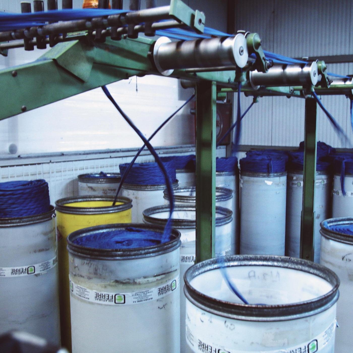 Cylindrical containers under a large green machine; blue threads are fed into the machine from each container.