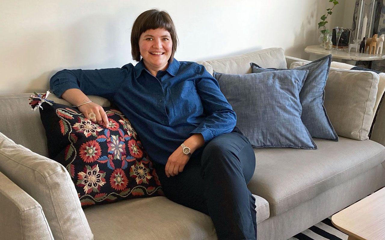 A smiling woman in blue clothes sitting on a grey sofa with cushions.