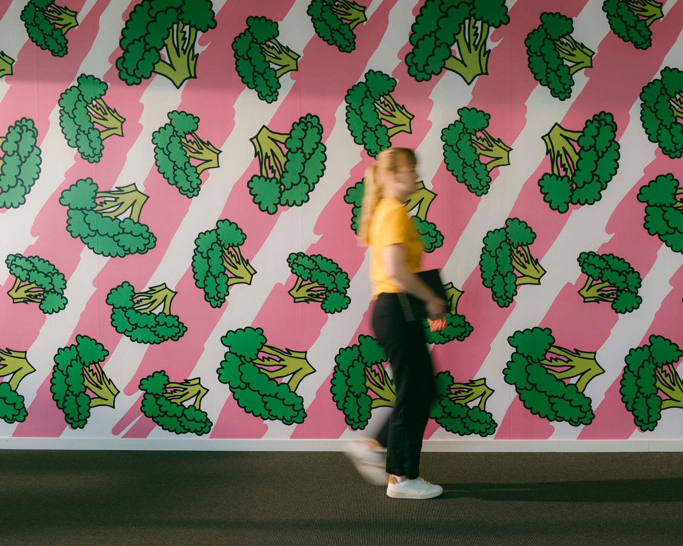 A woman walks in front of a wall decorated with a graphic design in pink and white stripes with broccoli.