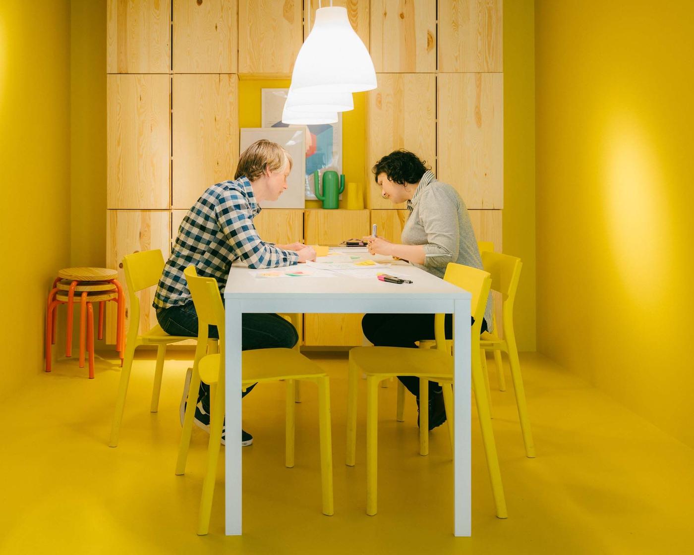 Two people sit in a yellow room at a white table. They work with lamps above them.
