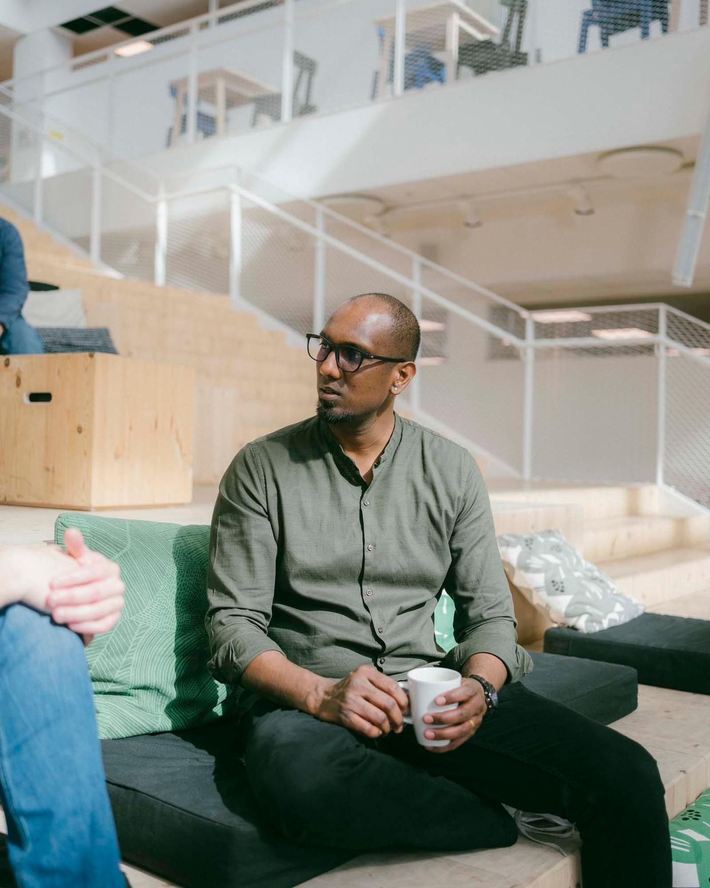 A man sits with a cup in his hands. He is talking to someone. There is a staircase in the background.
