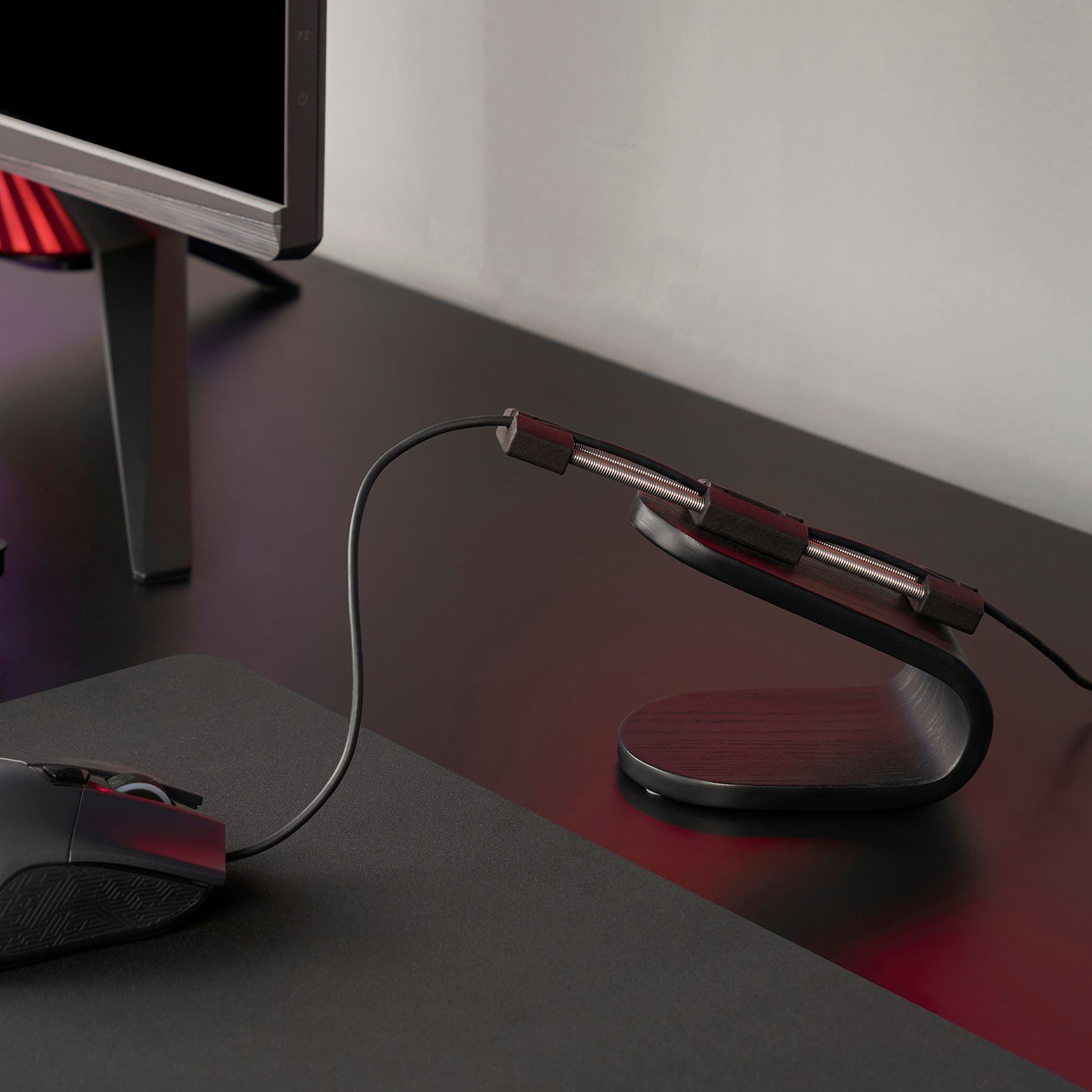 A black computer mouse on a grey mouse pad, connected to a small black table stand next to a computer screen.