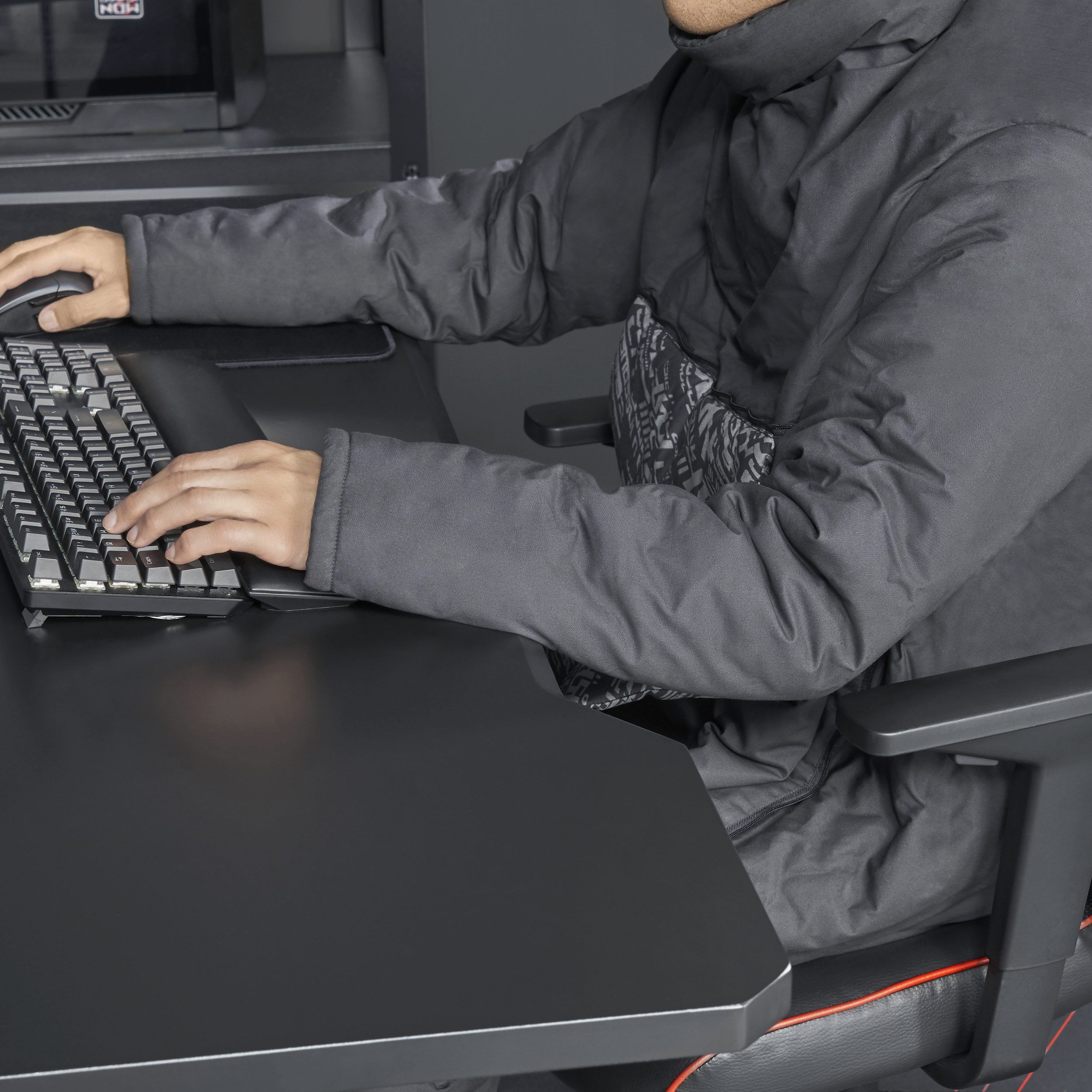 A person in a grey jacket sitting in a black office chair at a black desk working with a computer keyboard and a mouse.