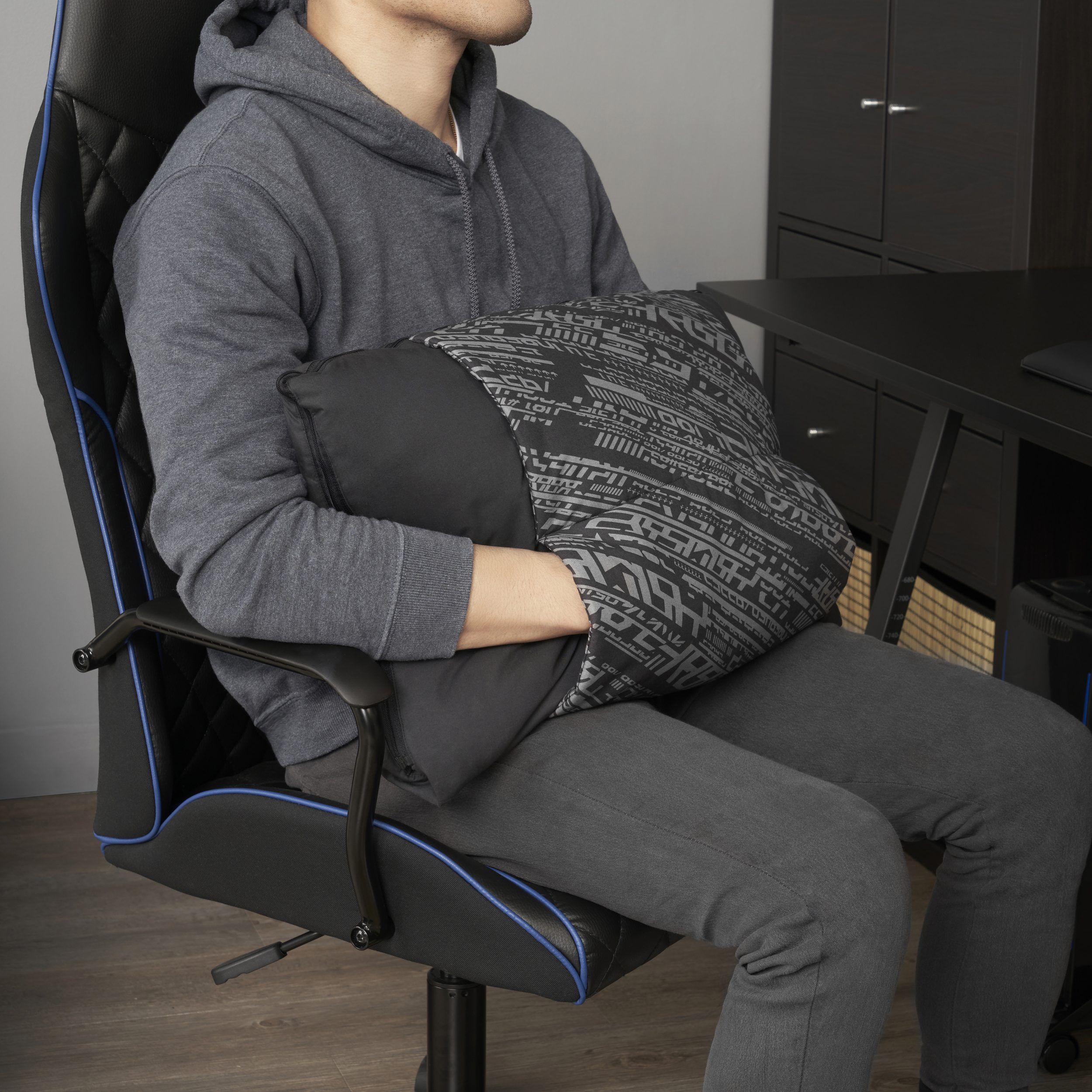 A man in a grey hoodie with his hands stuck inside a black patterned muff, sitting on a black office chair by a black desk.