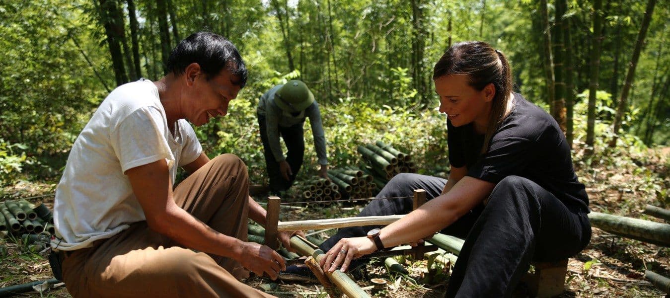 A man and a woman sawing bamboo in a forest, with another man in the background.