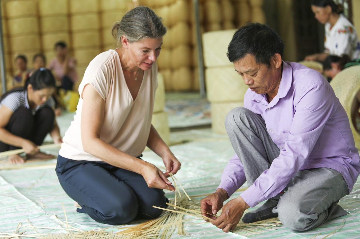 A woman and a man hand-weaving bamboo on a rug on the floor, with other workers in the background.