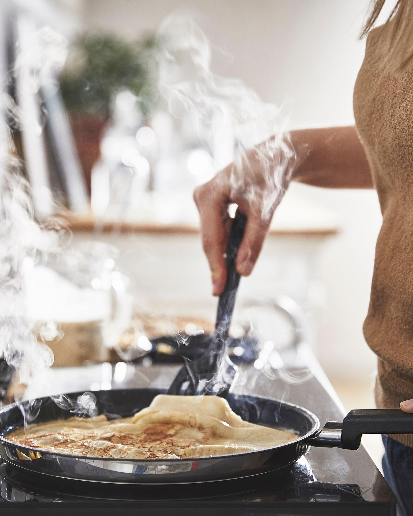The MIDDAGSMAT pan on the stove. In the pan there's a pancake. Someone is holding the pan and has a cooking utility underneath the pancake, holding the pancake up a little.